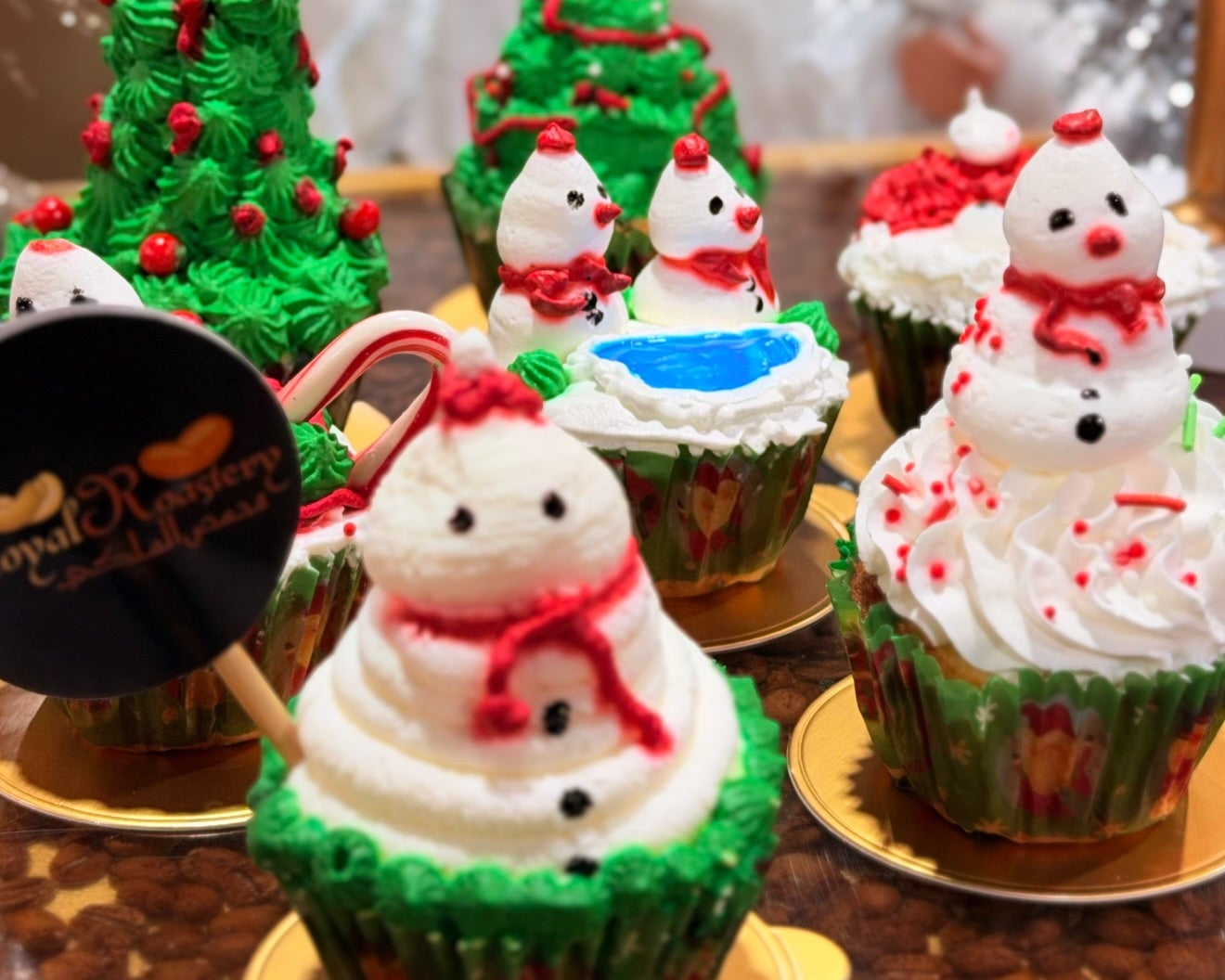 A festive collection of Christmas-themed cupcakes decorated with white frosting, green icing, red accents, and snowman faces, displayed on a table with a blurred Santa Claus figure in the background.