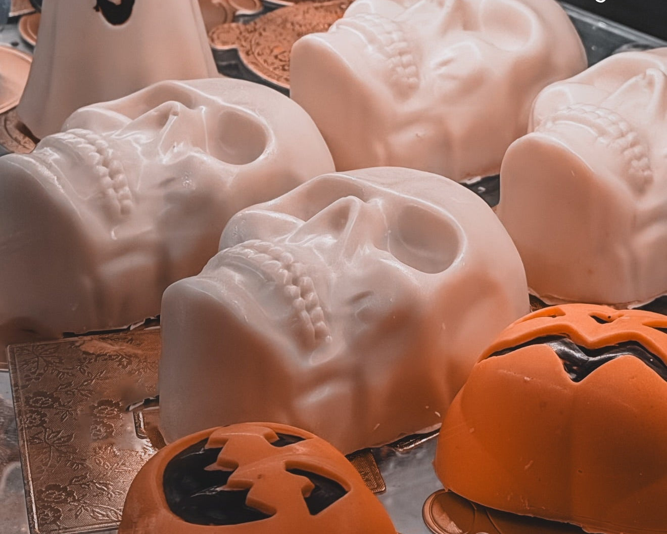 A display of Halloween gelato treats, including skull-shaped cones, a ghost cone with a smiling face, a pumpkin-shaped cone with black jack-o'-lantern details, and a blue brain-shaped cone, arranged on a glass display case.