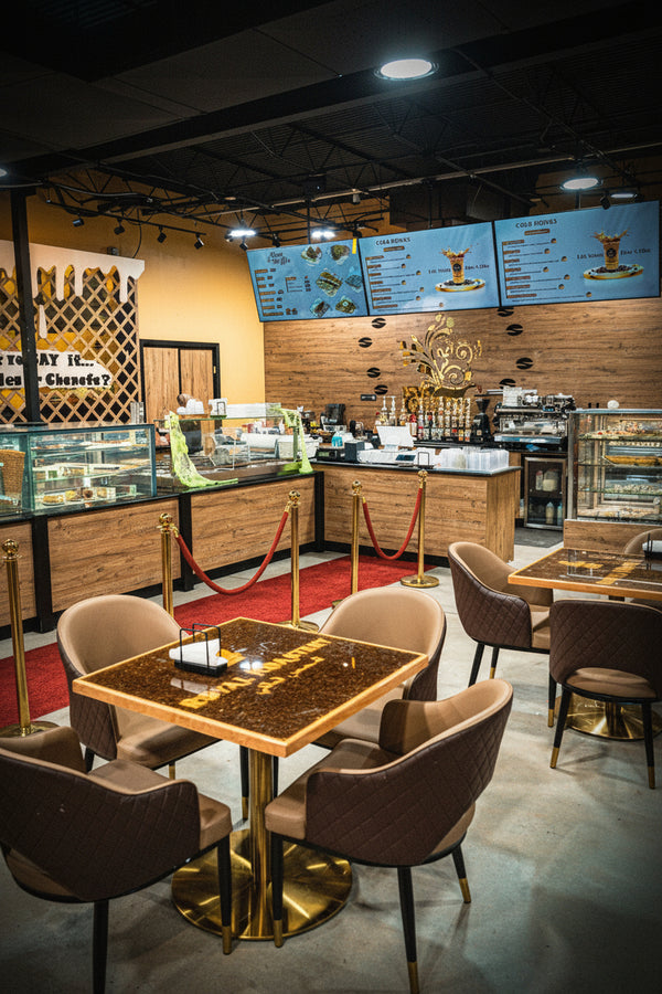 A modern cafe interior with multiple small square tables, brown quilted chairs, red velvet rope barriers, wood counter walls, and digital menu boards above the counter.