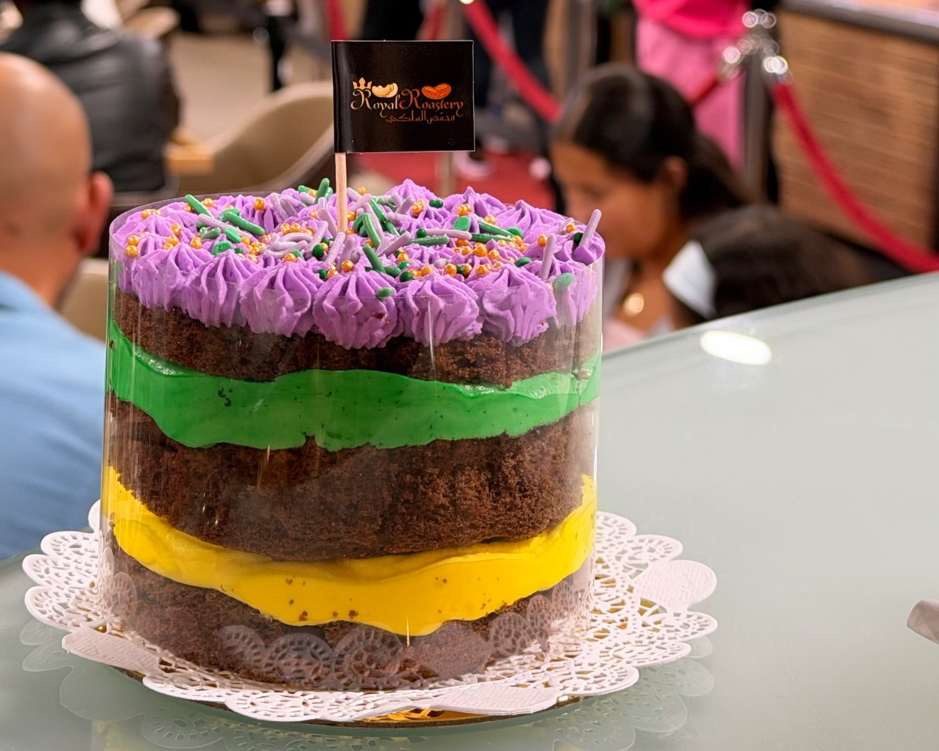 A colorful layered cake with purple, green, yellow, and brown layers sits on a table in a bakery or cafe, with blurred people and store interior in the background.